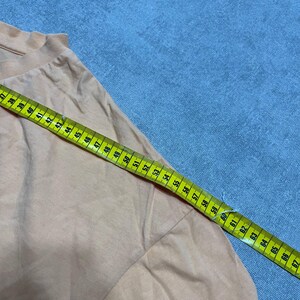 May include: A peach-colored garment with a ruffled neckline is measured with a yellow and green measuring tape. The tape shows measurements from 37 to 67 inches. The garment is on a blue surface.