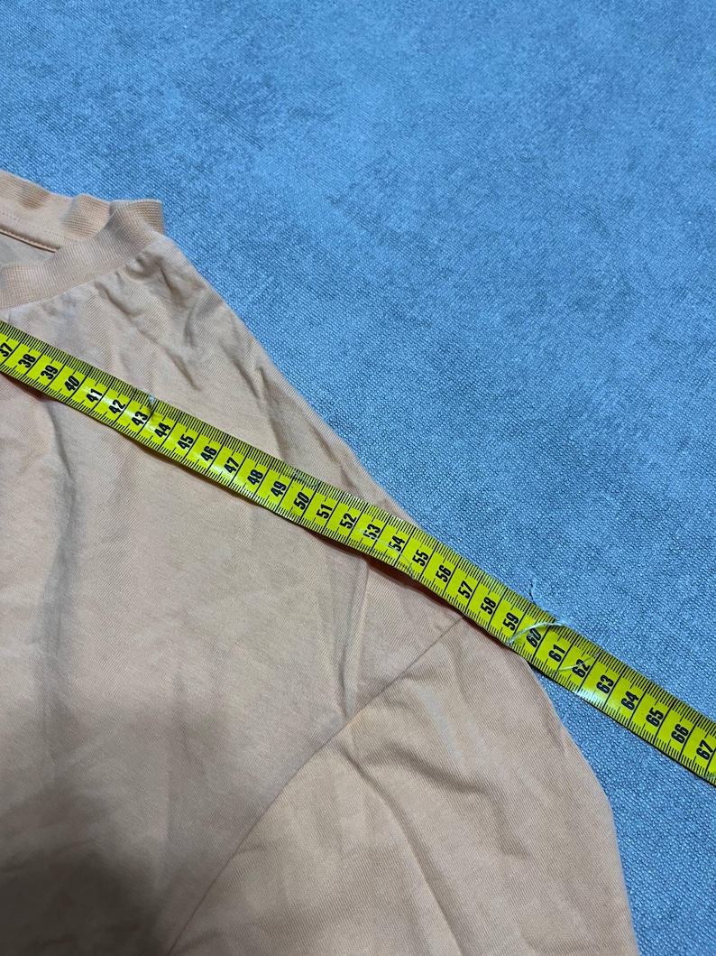 May include: A peach-colored garment with a ruffled neckline is measured with a yellow and green measuring tape. The tape shows measurements from 37 to 67 inches. The garment is on a blue surface.