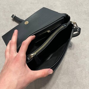 May include: Black crossbody bag with a flap closure and gold-toned hardware. The bag is open, revealing a zippered compartment and a smaller pocket. The strap is visible, and the interior appears to be lined with a dark fabric.