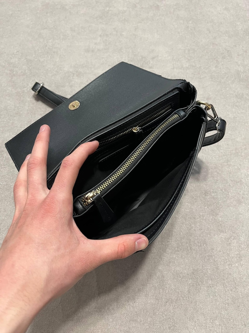 May include: Black crossbody bag with a flap closure and gold-toned hardware. The bag is open, revealing a zippered compartment and a smaller pocket. The strap is visible, and the interior appears to be lined with a dark fabric.