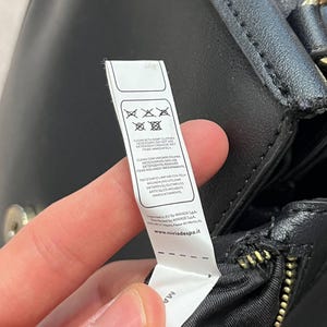 May include: A black leather handbag with a gold zipper and a white tag attached. The tag displays cleaning instructions and the website www.miriadespa.it.