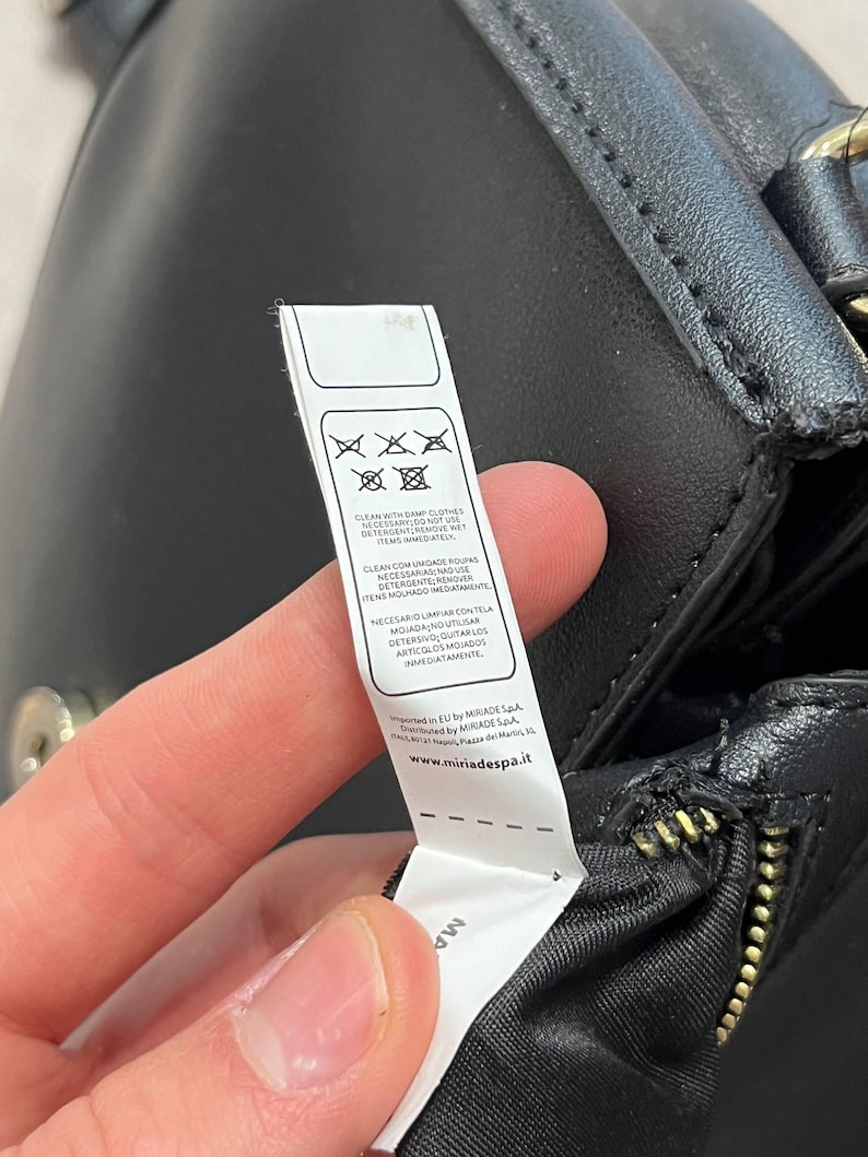 May include: A black leather handbag with a gold zipper and a white tag attached. The tag displays cleaning instructions and the website www.miriadespa.it.