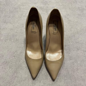Puede incluir: Un par de zapatos de tac&oacute;n alto de charol beige. Los zapatos tienen punta afilada y tac&oacute;n de aguja. El logotipo de Valentino es visible en la plantilla. Los zapatos est&aacute;n sobre un fondo neutro.
