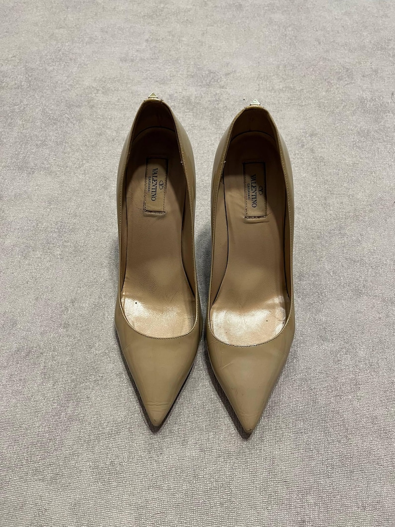 Puede incluir: Un par de zapatos de tac&oacute;n alto de charol beige. Los zapatos tienen punta afilada y tac&oacute;n de aguja. El logotipo de Valentino es visible en la plantilla. Los zapatos est&aacute;n sobre un fondo neutro.