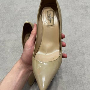 Puede incluir: Zapatos de tac&oacute;n beige de charol con punta. Los zapatos tienen un acabado brillante y un tac&oacute;n de aguja. El interior del zapato es de color beige claro. El logotipo de Valentino es visible en el interior.