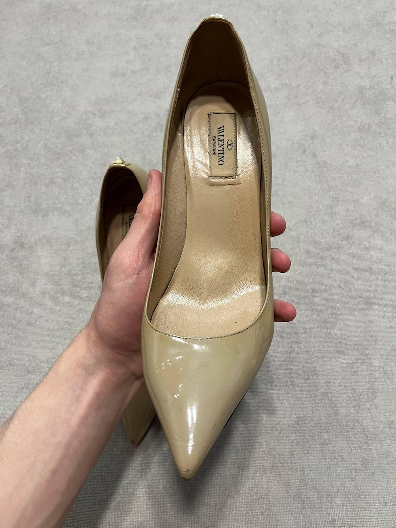 Puede incluir: Zapatos de tac&oacute;n beige de charol con punta. Los zapatos tienen un acabado brillante y un tac&oacute;n de aguja. El interior del zapato es de color beige claro. El logotipo de Valentino es visible en el interior.