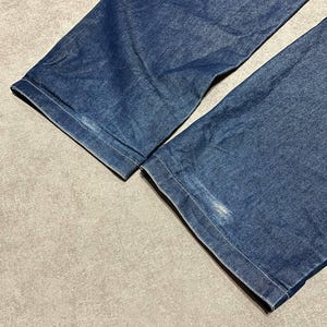 Puede incluir: Primer plano de dos perneras de pantal&oacute;n vaquero azul denim. Los vaqueros son de un lavado medio con costuras visibles a lo largo de los dobladillos. La tela muestra algo de desgaste, con algunas manchas m&aacute;s claras en el denim.