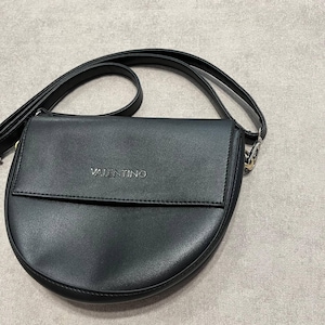 May include: Black crossbody bag with a curved bottom and a flap closure. The bag has a long, adjustable strap and the brand name "VALENTINO" is embossed on the front. The bag is made of a smooth, leather-like material.