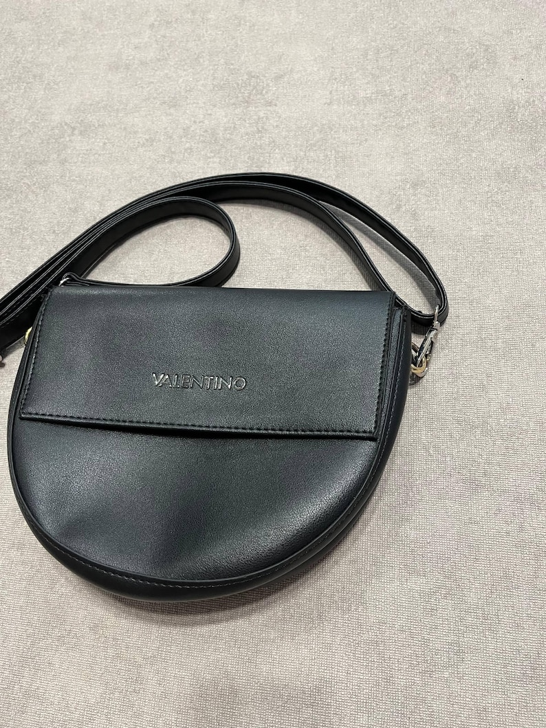 May include: Black crossbody bag with a curved bottom and a flap closure. The bag has a long, adjustable strap and the brand name "VALENTINO" is embossed on the front. The bag is made of a smooth, leather-like material.
