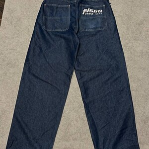 Puede incluir: Vaqueros de mezclilla azul oscuro con un dise&ntilde;o de pierna ancha. La parte trasera presenta dos bolsillos y una cinturilla blanca con el texto "FISGO JEANS" y "URBAN GEAR". Los vaqueros tienen un corte relajado.