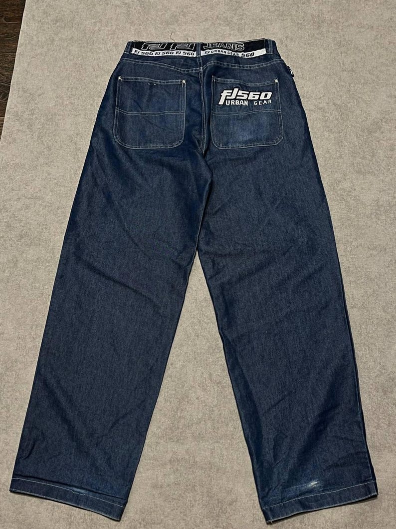 Puede incluir: Vaqueros de mezclilla azul oscuro con un dise&ntilde;o de pierna ancha. La parte trasera presenta dos bolsillos y una cinturilla blanca con el texto "FISGO JEANS" y "URBAN GEAR". Los vaqueros tienen un corte relajado.