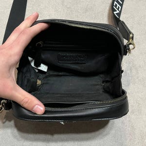 May include: Black crossbody bag with a zippered closure and a long strap. The strap has the word "VALEN" printed on it in white. The bag is open, revealing the interior. The bag has a small chain on the side.