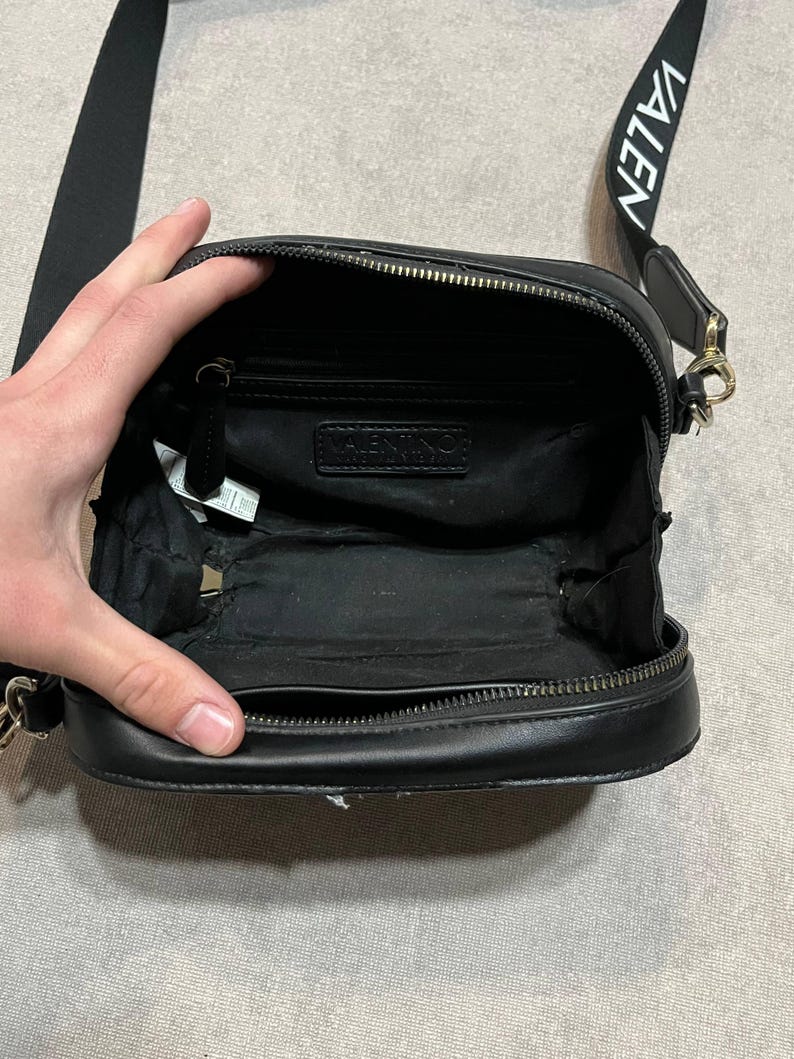 May include: Black crossbody bag with a zippered closure and a long strap. The strap has the word "VALEN" printed on it in white. The bag is open, revealing the interior. The bag has a small chain on the side.
