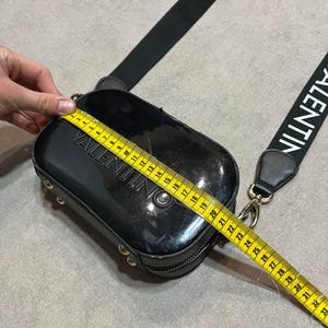 May include: Black rectangular crossbody bag with the brand name "VALENTINO" embossed on the front. The bag has a black strap with the brand name printed in white. The bag measures approximately 10 inches in length.
