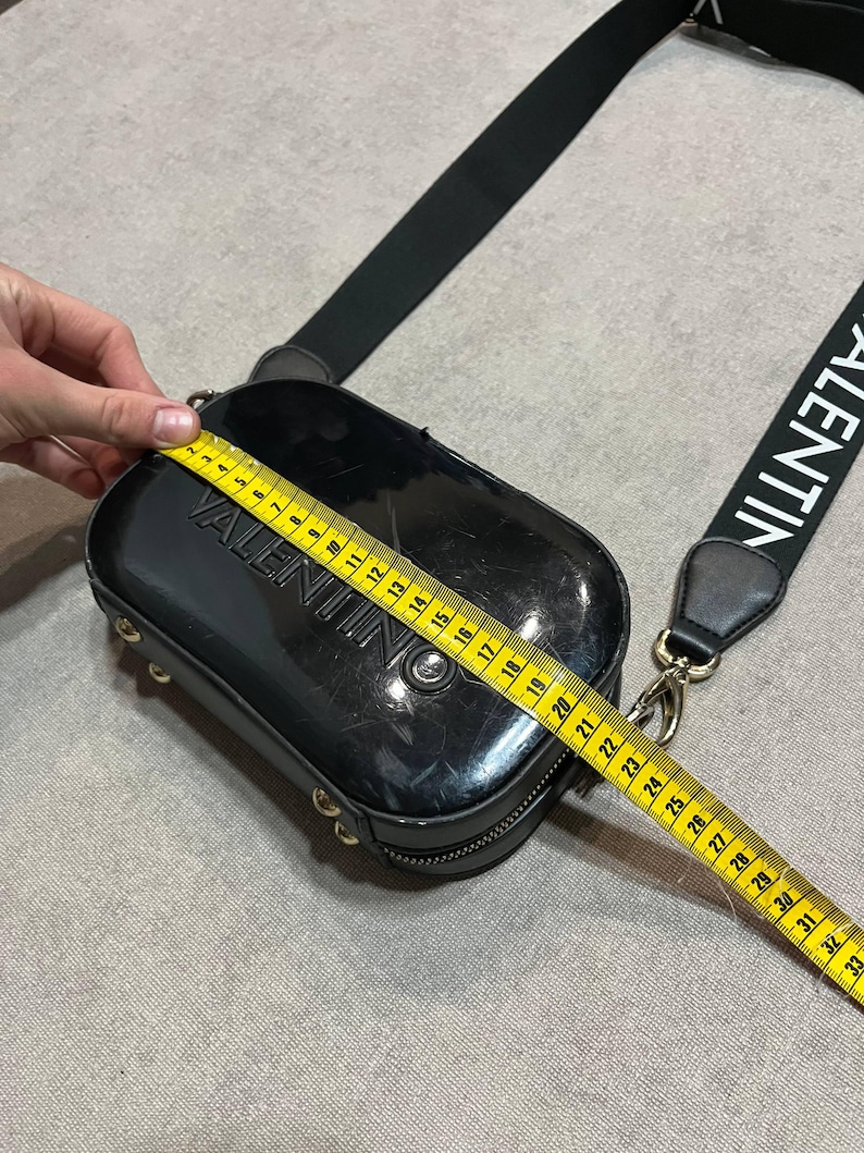 May include: Black rectangular crossbody bag with the brand name "VALENTINO" embossed on the front. The bag has a black strap with the brand name printed in white. The bag measures approximately 10 inches in length.