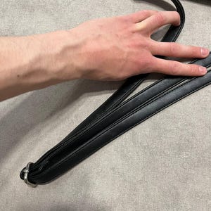 May include: Black leather shoulder bag with a silver-tone metal clasp and strap. The bag's strap is held by a hand. The bag is on a gray surface.