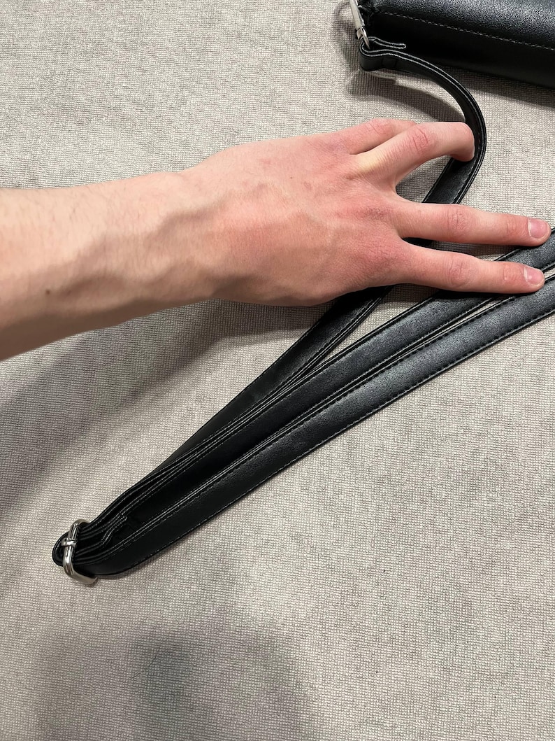 May include: Black leather shoulder bag with a silver-tone metal clasp and strap. The bag's strap is held by a hand. The bag is on a gray surface.