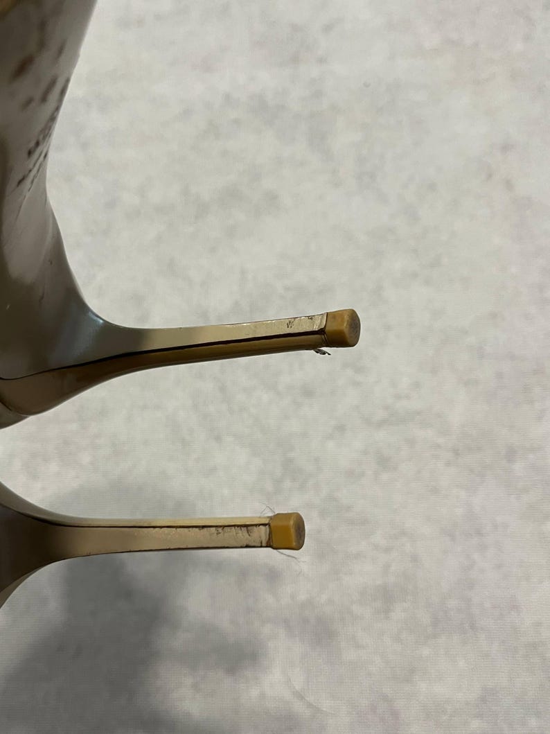 Puede incluir: Primer plano de dos zapatos de tac&oacute;n alto. Los tacones son de color beige claro con un dise&ntilde;o fino y de aguja. Las puntas de los tacones tienen una peque&ntilde;a tapa cuadrada de color marr&oacute;n claro. Los zapatos parecen estar hechos de un material liso.