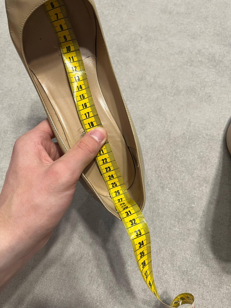Puede incluir: Un zapato plano beige de mujer se mide con una cinta m&eacute;trica amarilla. La cinta muestra una medida de aproximadamente 45 cm. El zapato tiene una punta redondeada y un tac&oacute;n bajo. El interior del zapato es de color beige claro.