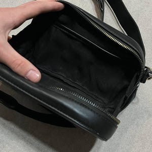 May include: Black crossbody bag with a zippered main compartment and a long, adjustable strap. The bag is open, revealing a black interior with a small pocket. The strap is black with white lettering. The bag is made of a smooth, leather-like material.