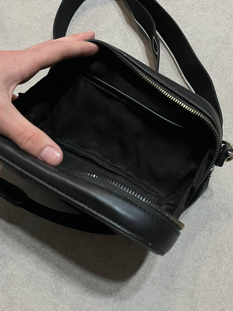 May include: Black crossbody bag with a zippered main compartment and a long, adjustable strap. The bag is open, revealing a black interior with a small pocket. The strap is black with white lettering. The bag is made of a smooth, leather-like material.