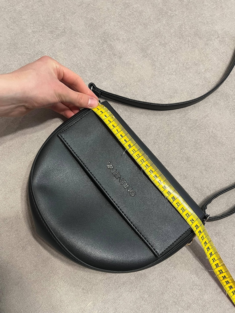 May include: Black Valentino crossbody bag with a flap closure and a long strap. The bag is measured with a tape measure, showing a length of approximately 14 inches. The word "VALENTINO" is embossed on the front.