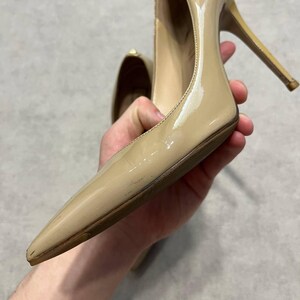 Puede incluir: Un par de tacones altos de charol beige con punta y tacones de aguja. Los zapatos tienen un acabado brillante y se sostienen en la mano de una persona. Los tacones miden aproximadamente 10 cm de alto.