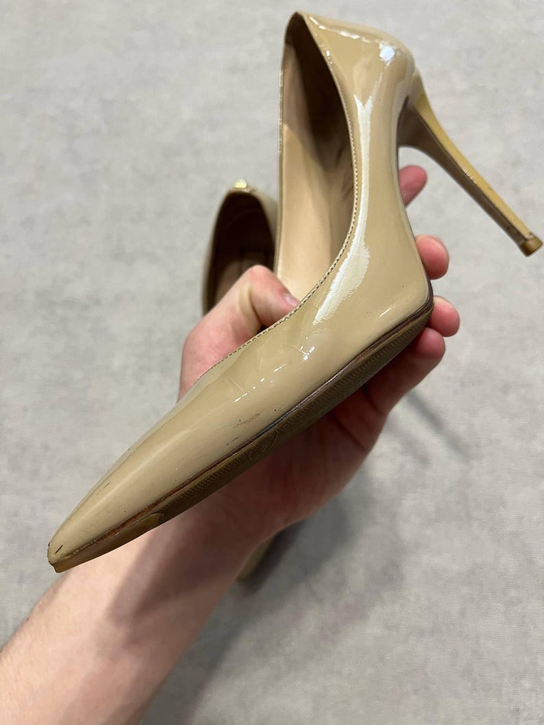 Puede incluir: Un par de tacones altos de charol beige con punta y tacones de aguja. Los zapatos tienen un acabado brillante y se sostienen en la mano de una persona. Los tacones miden aproximadamente 10 cm de alto.