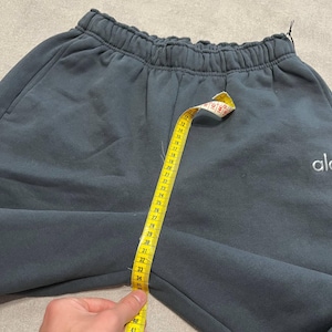 May include: Dark charcoal gray sweatpants with an elastic waistband and the text "alo" embroidered in white on the right side. A yellow measuring tape is placed vertically from the waistband to the crotch, showing a measurement of 23 inches.