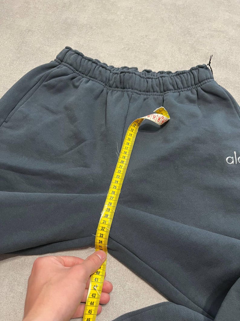 May include: Dark charcoal gray sweatpants with an elastic waistband and the text "alo" embroidered in white on the right side. A yellow measuring tape is placed vertically from the waistband to the crotch, showing a measurement of 23 inches.