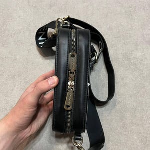 May include: Black rectangular crossbody bag with a gold zipper and hardware. The bag has a black strap with white text and a black adjustable shoulder strap. The bag is held by a person's hand.