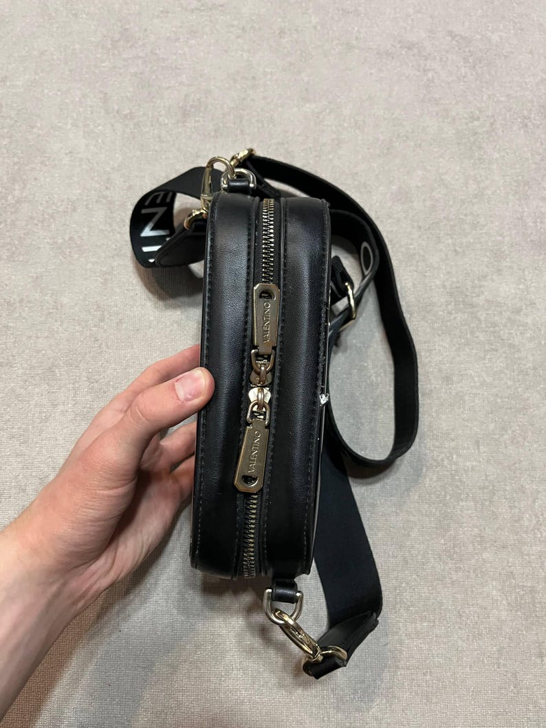 May include: Black rectangular crossbody bag with a gold zipper and hardware. The bag has a black strap with white text and a black adjustable shoulder strap. The bag is held by a person's hand.