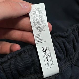 May include: A white rectangular garment care label attached to a dark charcoal fabric waistband. The label features printed text including company addresses in Indonesia, Turkey, and Korea, style number U5012RG, and recycling icons with the text quetairedemedeschets.fr.