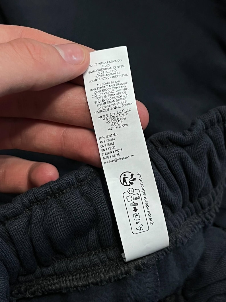 May include: A white rectangular garment care label attached to a dark charcoal fabric waistband. The label features printed text including company addresses in Indonesia, Turkey, and Korea, style number U5012RG, and recycling icons with the text quetairedemedeschets.fr.