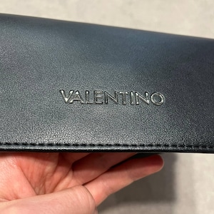 May include: A dark navy blue leather accessory with the embossed silver word "VALENTINO". The accessory has a textured surface and a stitched edge. The item is held in a hand.