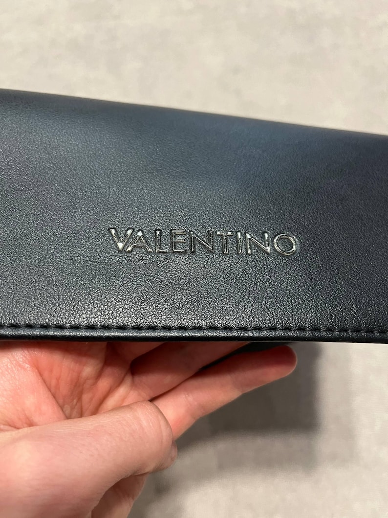 May include: A dark navy blue leather accessory with the embossed silver word "VALENTINO". The accessory has a textured surface and a stitched edge. The item is held in a hand.