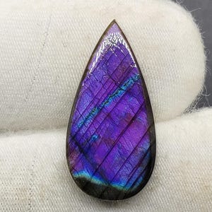 May include: A teardrop-shaped, polished labradorite gemstone. The stone displays a vibrant play of color, with deep purple and blue hues. The surface has a subtle, textured pattern, and the stone is set against a white background.