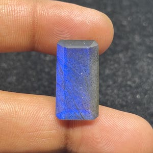 Peut inclure: Une pierre précieuse de labradorite rectangulaire avec une surface polie et facettée. La pierre présente un éclat bleu vif avec des inclusions grises et noires. La pierre est tenue entre deux doigts, mettant en valeur sa taille et sa couleur.
