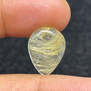 以下が含まれることがあります： 金色の針状のインクルージョンが入った、透明な涙型の宝石。 宝石は2本の指の間に挟まれ、研磨された表面と内部の模様が示されています。 石の透明度により光が透過し、金色のストランドが強調されています。