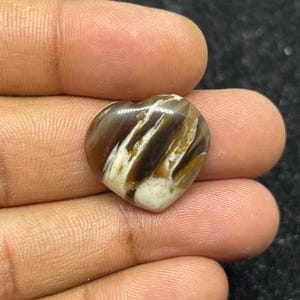 May include: A heart-shaped gemstone with a polished surface. The stone features a mix of brown, white, and tan colors, with a swirling pattern. The gemstone is held in a person's hand, showcasing its size and shape.