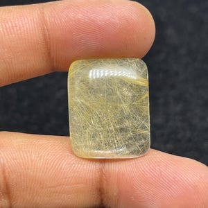 以下が含まれることがあります： 金色の針状のインクルージョンが入った、長方形の透明な宝石。 磨かれた表面と内部の複雑な模様が、2本の指の間に挟まれて見えています。 宝石の透明度により光が通り、金色のストランドが強調されています。