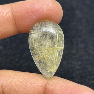 以下が含まれることがあります： 金色の針状のインクルージョンが入った、透明な涙型の宝石。 宝石は2本の指の間に挟まれ、研磨された表面と内部の模様が示されています。 石の透明度により光が透過し、金のストランドが強調されています。