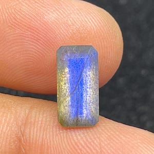 Peut inclure: Une pierre précieuse rectangulaire avec un effet irisé bleu et or. La gemme est tenue entre les doigts, mettant en valeur sa surface polie et ses jeux de couleurs. La pierre est de forme rectangulaire et semble être une labradorite.