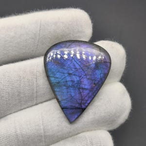 Peut inclure: Une pierre précieuse en forme de goutte d'eau, présentant un mélange captivant de teintes bleues et violettes. La surface polie révèle des motifs complexes et un léger scintillement. La pierre est tenue dans un gant blanc, soulignant ses couleurs vives et sa texture unique.