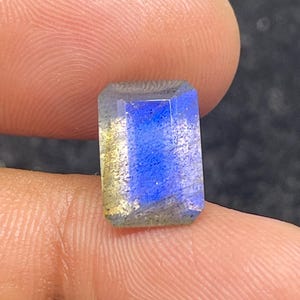 Peut inclure: Une pierre précieuse de coupe rectangulaire avec un scintillement irisé bleu et jaune. La gemme est tenue entre deux doigts, mettant en valeur ses facettes polies et son jeu de couleurs. Les bords de la pierre sont bien définis.
