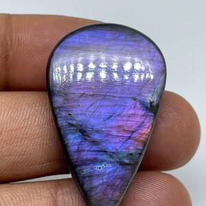 May include: A polished, teardrop-shaped labradorite gemstone. The stone exhibits a shimmering play of colour, with shades of purple, blue, and pink. The gemstone is held against a plain background, showcasing its iridescent qualities.