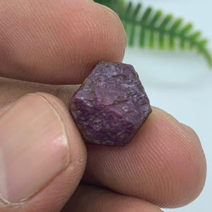 May include: A small, raw, hexagonal-shaped ruby gemstone with a deep, saturated purplish-red colour and a textured, matte surface. The stone displays natural crystalline facets and irregular edges, showcasing its unpolished, organic appearance.