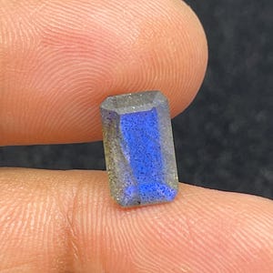 Peut inclure: Une pierre précieuse de coupe rectangulaire avec un éclat bleu vif, tenue entre les doigts. La gemme a un corps gris et clair avec une irisation bleue saisissante. Les facettes de la pierre captent la lumière, mettant en valeur sa surface polie.