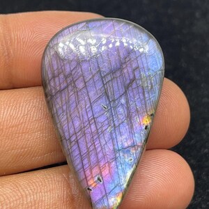Peut inclure: Une pierre précieuse en forme de goutte polie. La pierre présente un jeu de couleurs éclatant, avec des teintes dominantes de violet et de bleu, et des touches de jaune et d'orange. La surface est ornée d'un réseau de fines lignes.