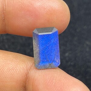 Peut inclure: Une pierre précieuse rectangulaire et facettée, d'un bleu vif et d'un dessus gris foncé. La pierre est tenue entre deux doigts, mettant en valeur sa surface polie et ses motifs internes. La gemme mesure environ 1,9 cm de long.